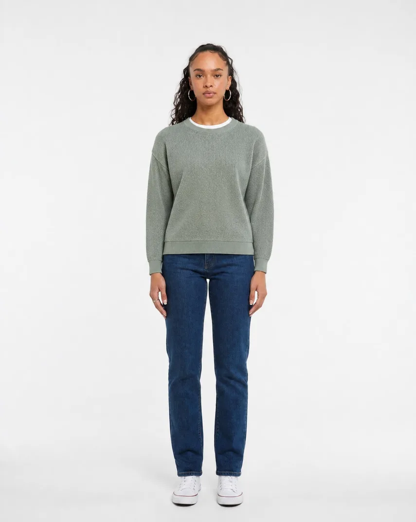 Model in a sage green coarse-gauge knit sweater with ribbed cuffs and hem over mid-blue jeans, front view against a white studio background